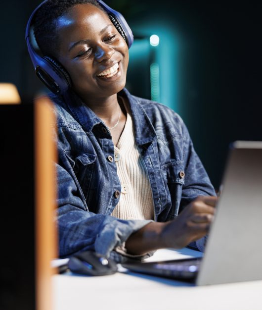Joyful African American blogger working from home, wearing wireless headset, typing on her laptop. Cheerful black woman multitasks, streaming movies and attending video calls with clients.